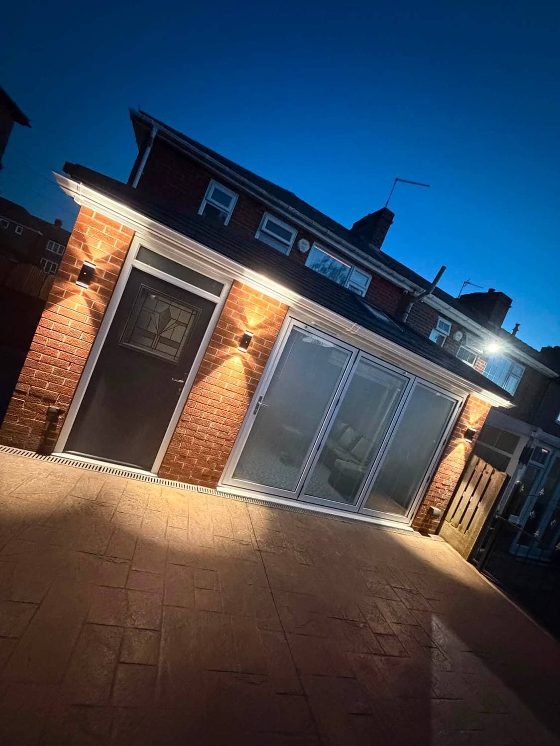 A rear extension at dusk with wide sliding doors lit from inside and warm uplighters on the brickwork